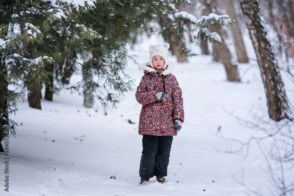 Smiling girl with white fur hat like a cat. Winter snowy background and gteen trees