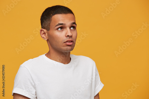 Attractive young man looks aside up puzzled, trying to remember something important, thoughtful. Saw something fascinating in the sky. Wears white casual t-shirt, on a yellow background