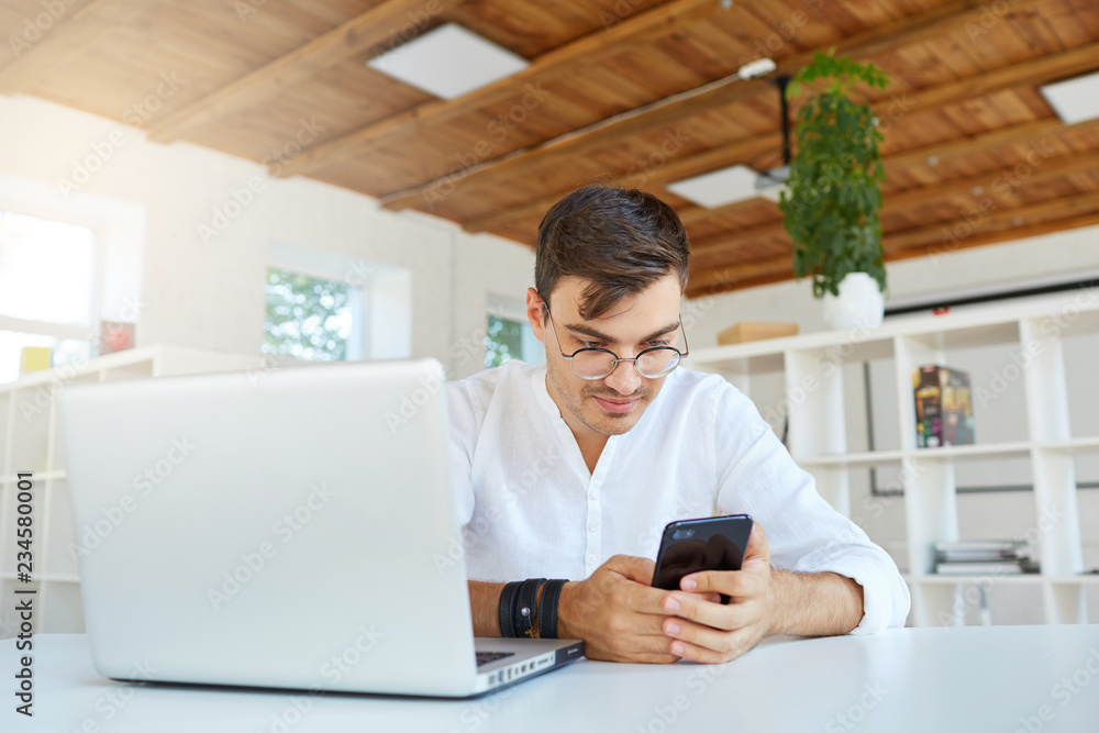 Portrait of serious handsome young businessman wears white shirt and glasses using laptop and smartphone sitting at the table in office Portrait of serious handsome young businessman wears white shirt and glasses using laptop and smartphone sitting at the table in office