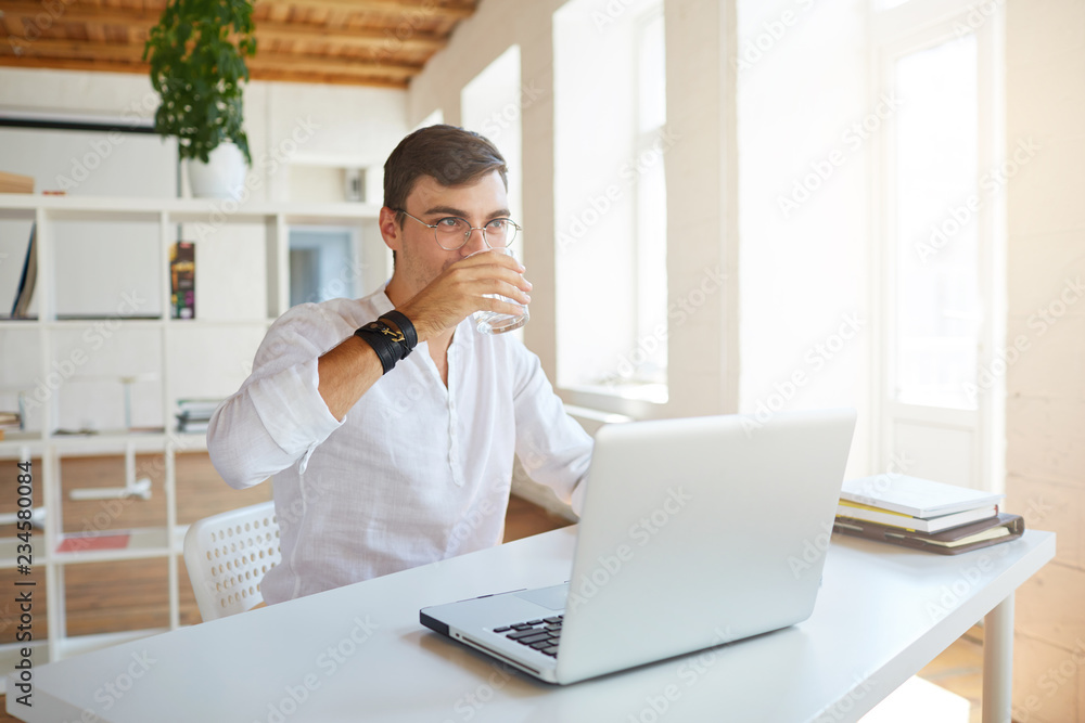 Closeup of attractive young businessman wears white shirt and spectacles working with laptop and drinking water at the table in office Closeup of attractive young businessman wears white shirt and spectacles working with laptop and drinking water at the table in office