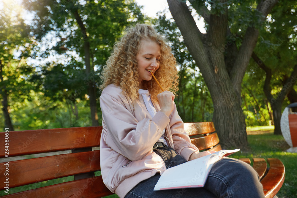 Student, beautiful blonde-hair girl sits on a park bench, with a notepad, looks on her notes, thinks about task, trying to find a solution, holding a pencil in the mouth and smiling, park background Student, beautiful blonde-hair girl sits on a park bench, with a notepad, looks on her notes, thinks about task, trying to find a solution, holding a pencil in the mouth and smiling, park background