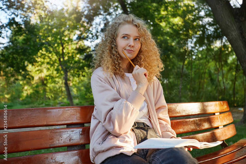 A young beautiful girl blonde sits on a park bench, with headphones, notes, a notepad, looks away, thinks about something, trying to find a solution, holding a pencil near the mouth, park background A young beautiful girl blonde sits on a park bench, with headphones, notes, a notepad, looks away, thinks about something, trying to find a solution, holding a pencil near the mouth, park background