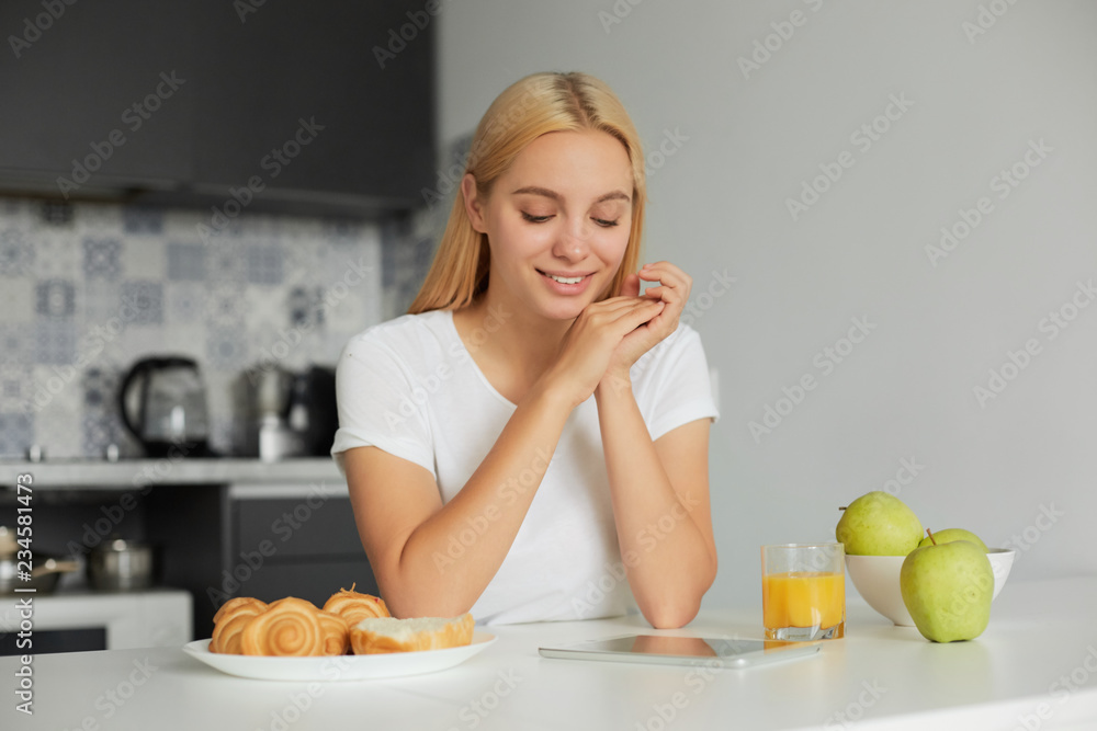 Young blonde girl sits at the kitchen table in the morning, smiling, looks at tablet black screen, on the table -a glass of juice, apples, buns, dressed in a domestic white T-shirt, kitchen background