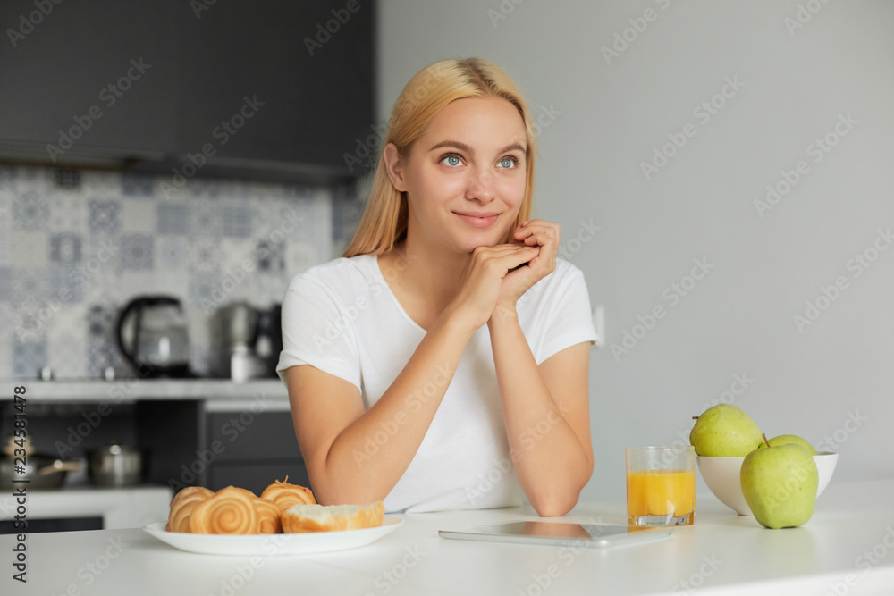 Photo of young blonde girl sits at the kitchen table in the morning, smiling, looks glad to the window, on the table -a glass of juice, apples, buns, dressed in a domestic T-shirt, kitchen background Photo of young blonde girl sits at the kitchen table in the morning, smiling, looks glad to the window, on the table -a glass of juice, apples, buns, dressed in a domestic T-shirt, kitchen background