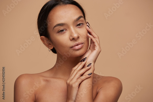 Charming mixed race girl, looking gently and tenderly like in the mirror after a face care mask, head slightly turned and tilted, touching her face, over beige background