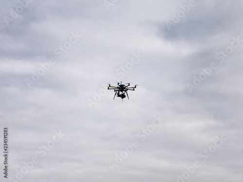 Large home made drone flying with a mirrorless camera hanging from it against acloudy, overcast sky background