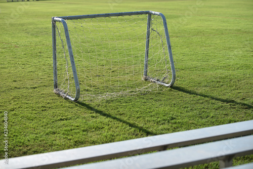 A small white aluminum goal with white net for kid’s football placed on a school pitch and stadium seatings in the afternoon waiting for a team to practice.