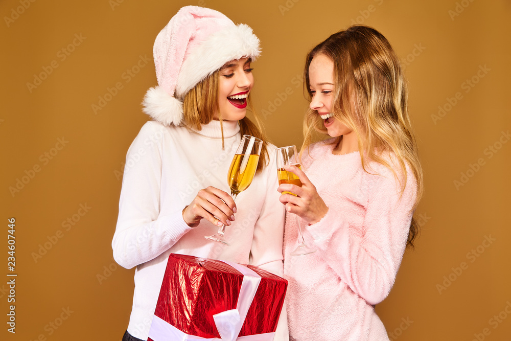 Christmas,x-mas, concept.Two smiling beautiful women in white winter sweaters.Girls posing on golden background.Models with big gift box drinking champagne in glasses.Having fun, celebrating New Year Christmas,x-mas, concept.Two smiling beautiful women in white winter sweaters.Girls posing on golden background.Models with big gift box drinking champagne in glasses.Having fun, celebrating New Year