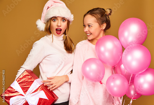 Christmas,x-mas, concept.Two smiling beautiful women in white winter sweaters.Girls posing on golden background.Models with big gift box and pink balloons.Having fun,ready for celebration new year