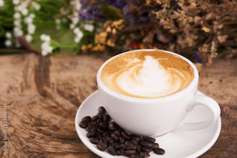 Obraz premium Cappuccino or latte art coffee. Free hand pouring some foam of milk on top. A white ceramic cup on the wood table with some coffee beans. The best food and drink in the world. Fresh energy everyday.