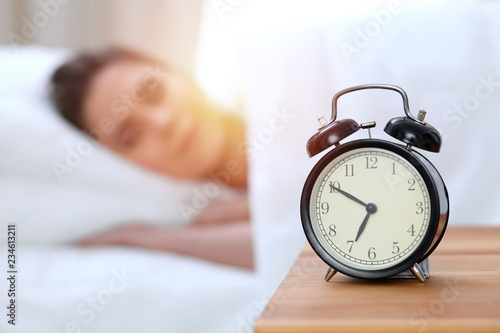 Alarm clock opposite of beautiful young woman sleeping while lying in bed comfortably and blissfully. Happy waking up early for a job