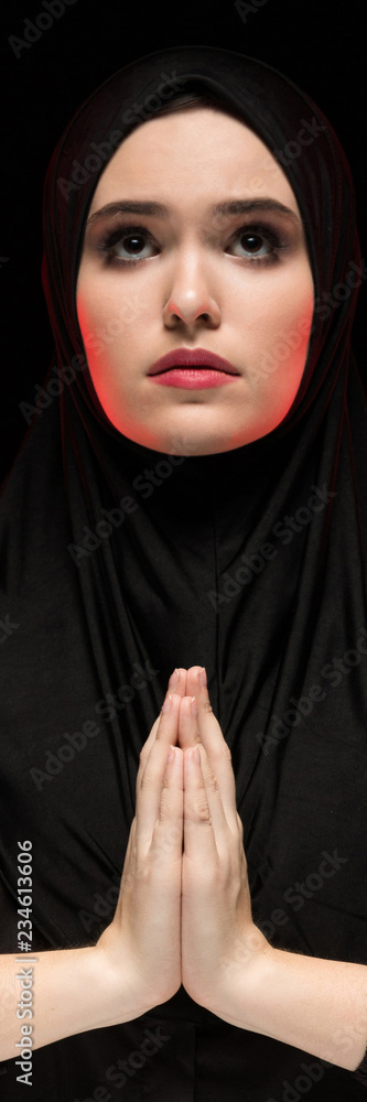 Portrait of beautiful serious young muslim woman wearing black hijab with hands near her face as praying concept on black background Portrait of beautiful serious young muslim woman wearing black hijab with hands near her face as praying concept on black background