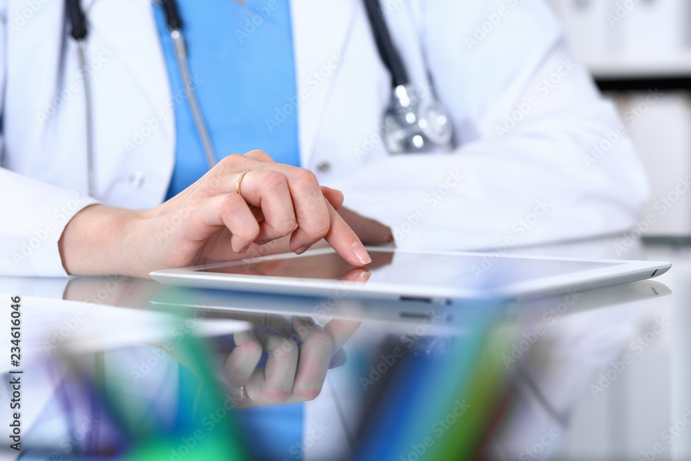 Woman doctor using tablet computer while sitting at the desk in hospital office, closeup. Healthcare, insurance and medicine concept Woman doctor using tablet computer while sitting at the desk in hospital office, closeup. Healthcare, insurance and medicine concept