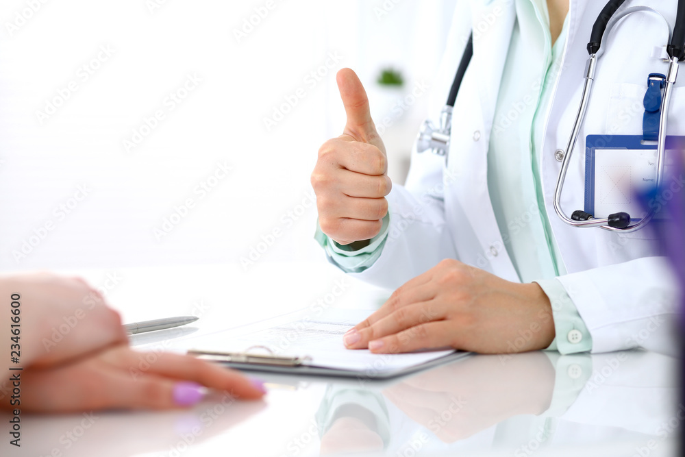 Doctor showing Ok sign with thumb up to patient while sitting at the desk in hospital office, close-up of human hands. Medicine and health care concept Doctor showing Ok sign with thumb up to patient while sitting at the desk in hospital office, close-up of human hands. Medicine and health care concept