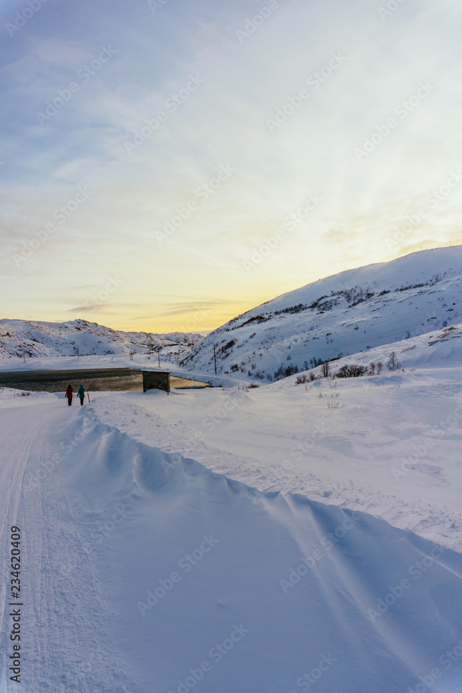 two people are walking in the distance along a snowy winter road in the village beyond the Arctic Circle to meet bright sunset light and a small sea bay