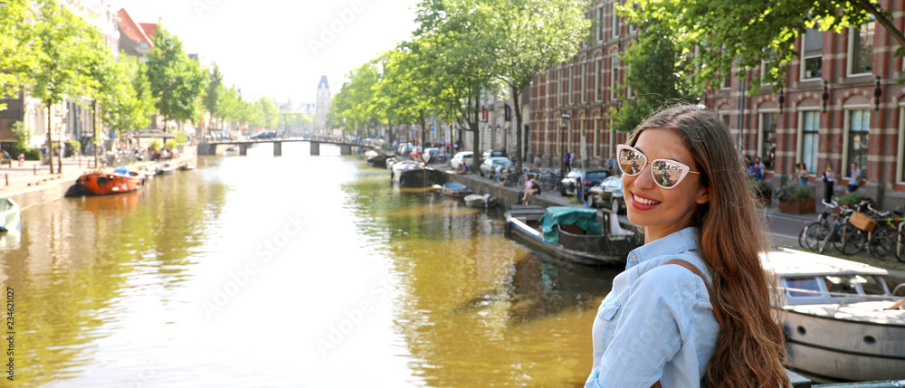 Naklejka premium Portret dziewczyny podróżnik z okulary i plecak korzystających z miasta Amsterdam. Panoramiczny widok banner młodej kobiety, patrząc do kamery na kanale Amsterdam, Holandia, Europa.