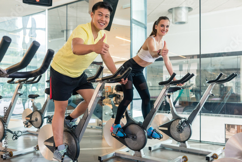 Fototapeta Naklejka Na Ścianę i Meble -  Young cheerful couple showing thumbs up while looking at camera during indoor cycling at the gym