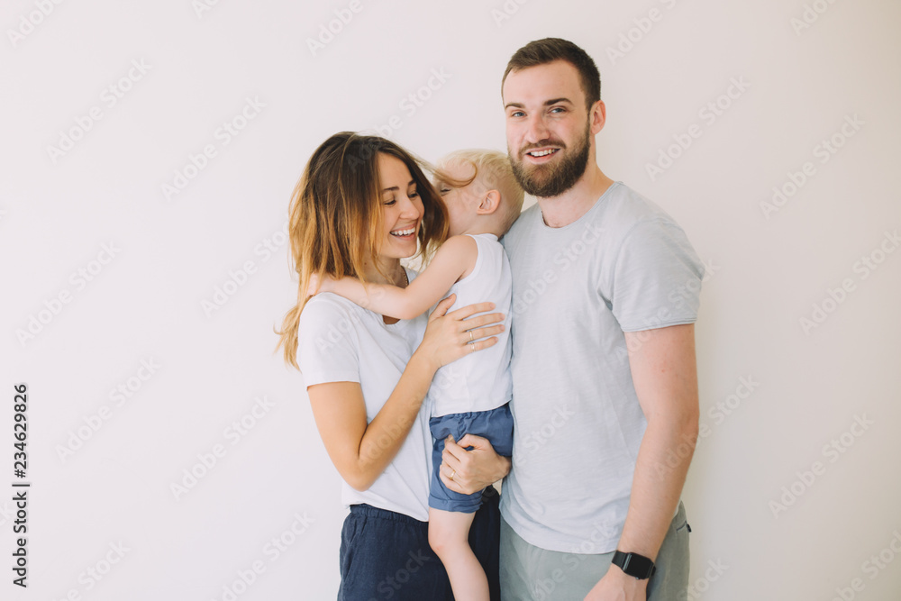 Happy young family with little son standing together in casuals near the empty wall - indoors .. - obrazy, fototapety, plakaty 