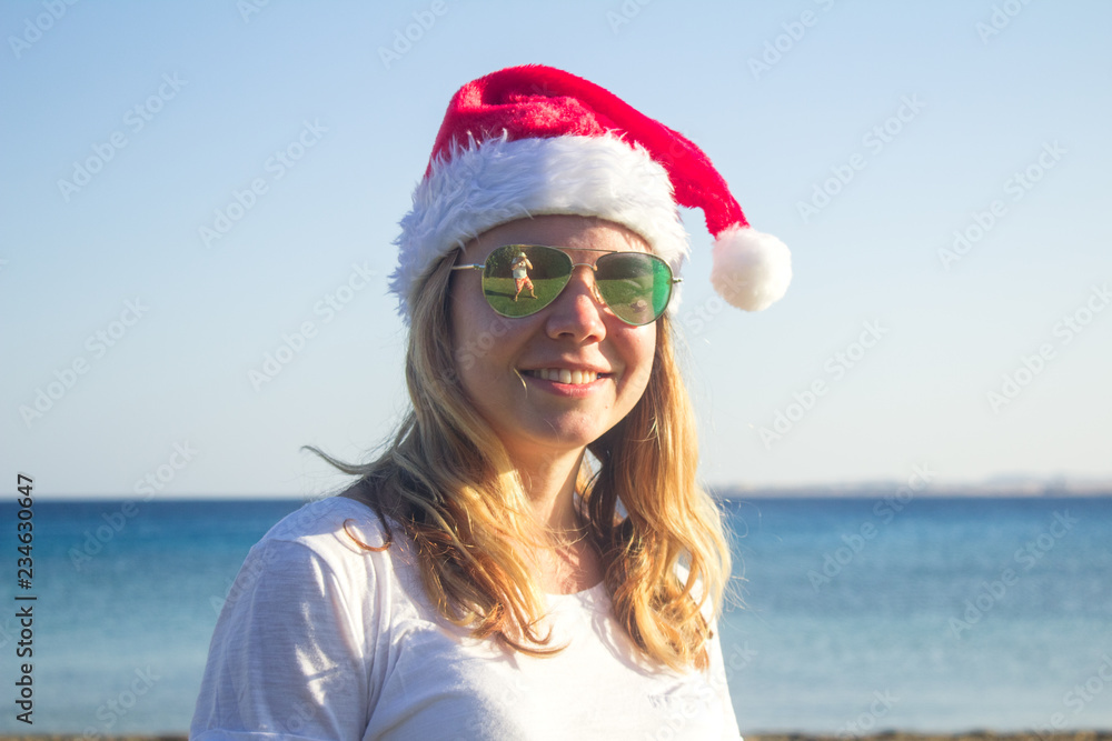 Obraz premium Portrait of a pretty young woman in Santa Claus hat and sunglasses on a sunny beach. Toned.
