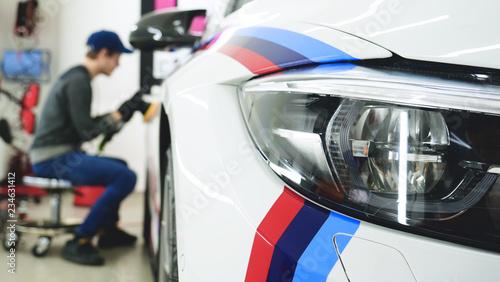 A man in a special suit polishes a gray car body, a tool for polishing cars, into a workshop. Concept of: Car service, Car wash, Polishing, Ceramics, Miscellaneous tools.