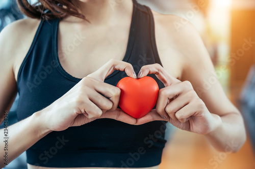 Asian sport woman holding red heart in fitness or gym center. Medical cadio heart strength training lifestyle. Pretty female sport girl workout exercise. Red massage ball in hand.