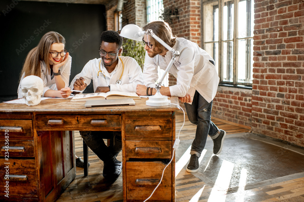 Group of a young multi ethnicity physicians or medical students in uniform working with book and drawings on the table in the office or classroom