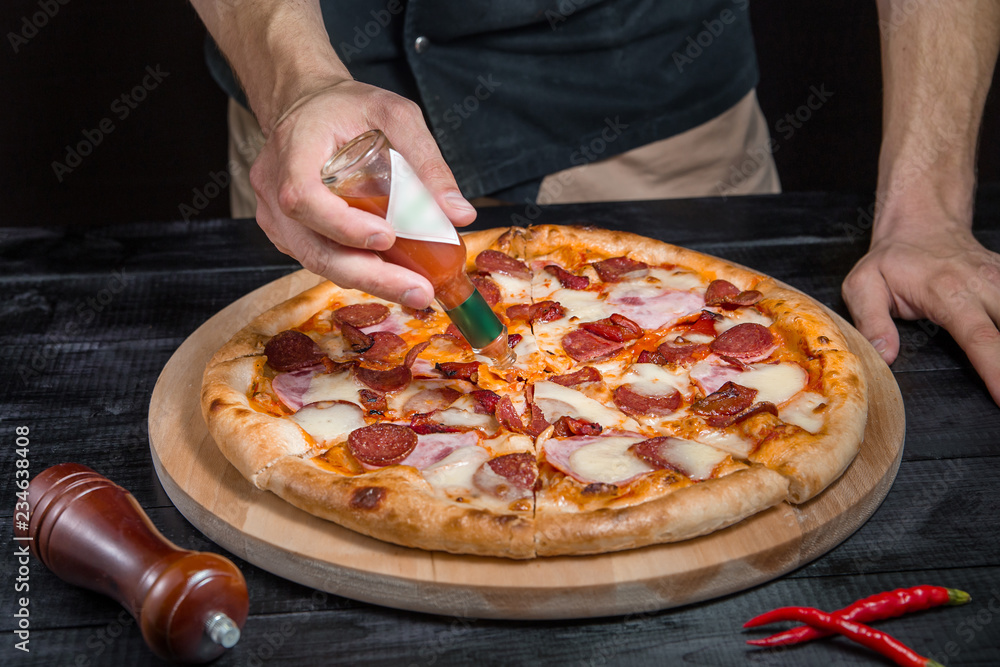 Cutting spicy pizza with salami, meat, ham, sausage, peppers, tomato, cheese with a pizza cutter on a wooden board on a black background