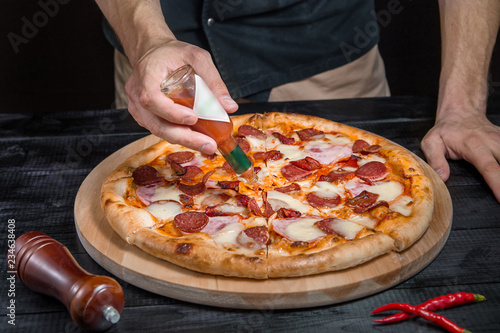 Cutting spicy pizza with salami, meat, ham, sausage, peppers, tomato, cheese with a pizza cutter on a wooden board on a black background