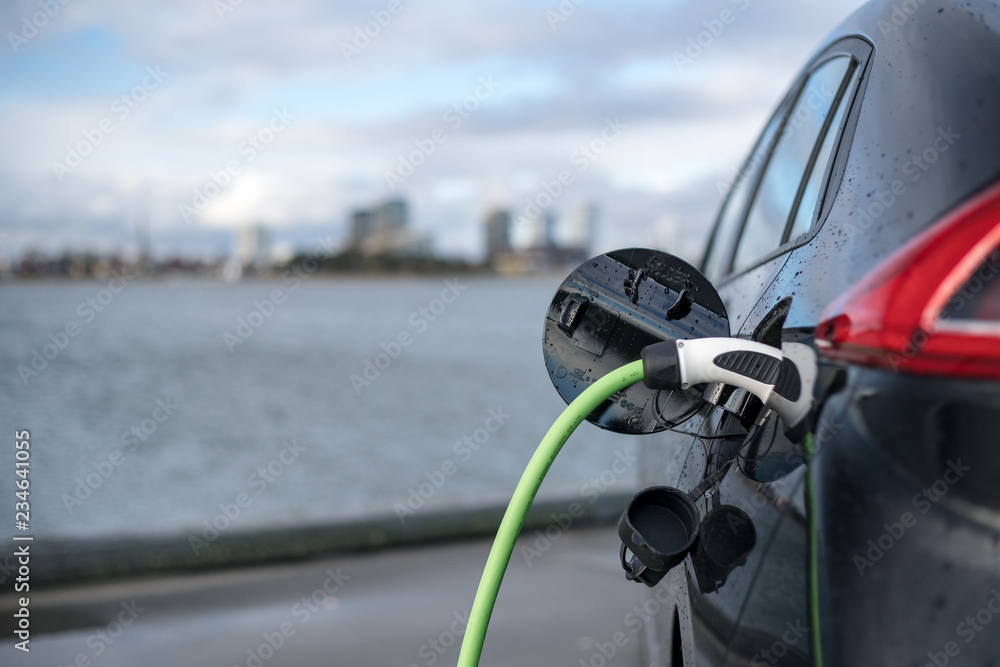 Charging an electric car, with the city and water in the background, green charging cable and black car. copenhagen