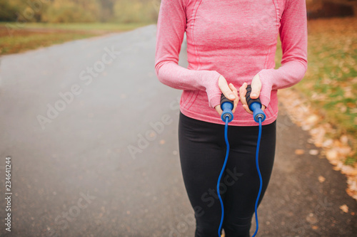 Cut view of well-buil young woman stand and holds blue jump rope in hands. She has rest. Woman is alone on road.