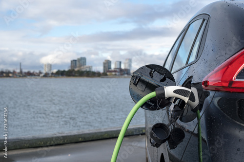 Charging an electric car, with the city and water in the background, green charging cable and black car. copenhagen