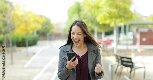 Front view of an excited woman walking towards camera, checking phone and celebrating good news in a park