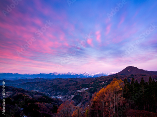 峠の朝焼けで青い空に、雲と遠くの山脈も紅く染ま　りる、手前には唐松と集落が見える。 photo