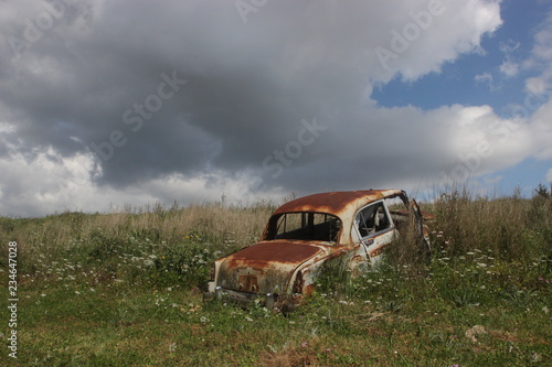 Abandoned old rusty vintage car on hill at summer with clouds at sky inside flowers green plants