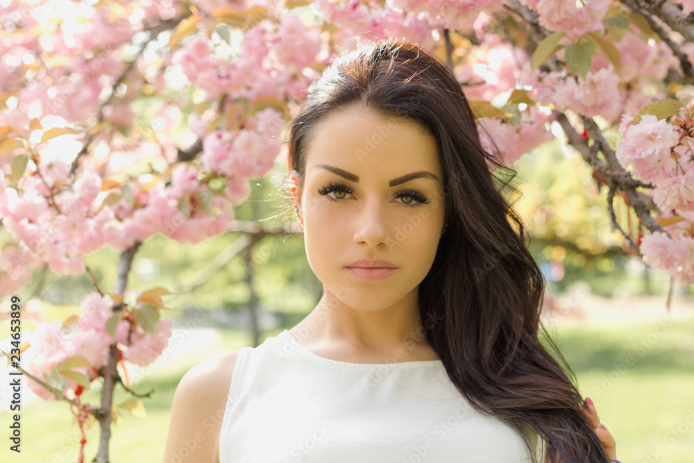 Attractive brunette woman in light dress looking at camera while posing near flowering tree Attractive brunette woman in light dress looking at camera while posing near flowering tree