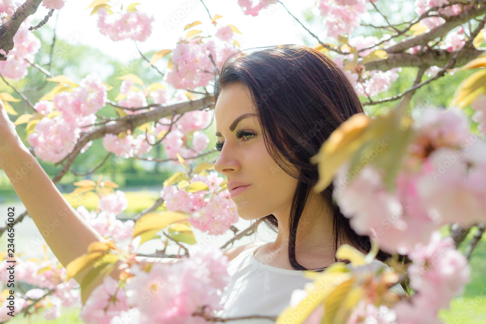 Fototapeta premium Attractive brunette woman in light dress looking at camera while posing near flowering tree