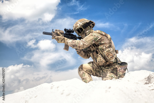 Man in military camouflage uniform with service rifle replica, standing on top of sand dune with cloudy sky on background, imitating U.S. army special forces shooter during airsoft war games in desert © Getmilitaryphotos