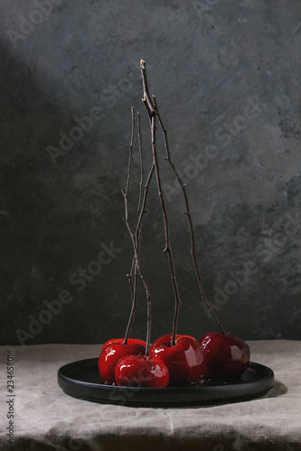 Red caramel apples sweet autumn or Christmas dessert served with branches in black ceramic plate on linen table cloth with grey wall at background. Dark atmospheric mood © Natasha Breen