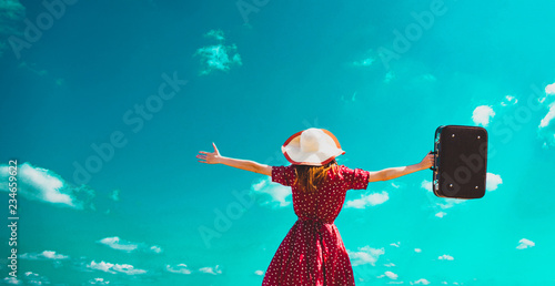 young girl in a hat and red dress with open arms holds a suitcase against the blue sky background