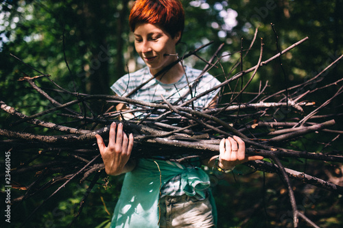 Red-haired hipster girl collects firewood on the background of the forest