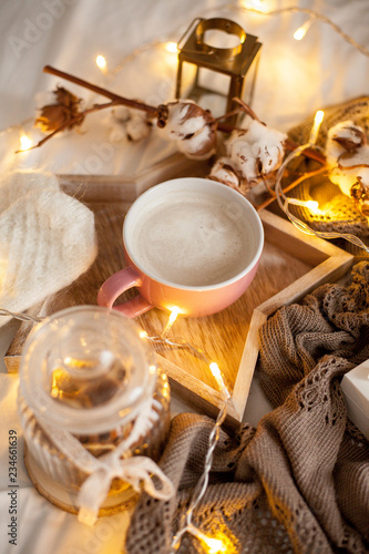 Mug of hot cappuccino on a wooden tray is on the bed. Cozy decor. Breakfast. Mug, plaid, cotton, candle. Gift box and knitted mittens. Christmas lights. Holidays. Christmas. Autumn. Winter.