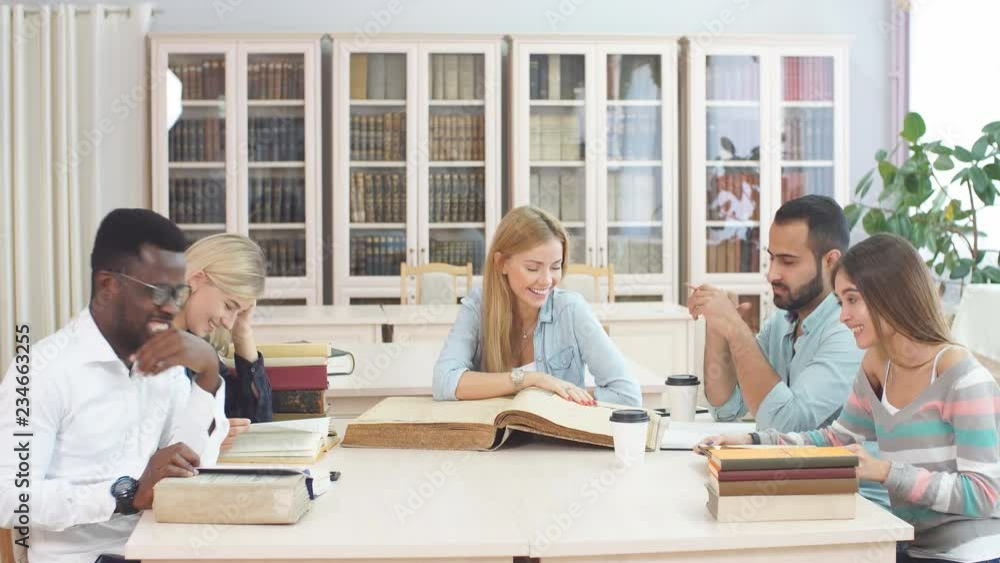 Multiethnic young people sitting at table reading big old reference book for preparing scientific report. Group of young students doing together homework at library. Multiethnic young people sitting at table reading big old reference book for preparing scientific report. Group of young students doing together homework at library.