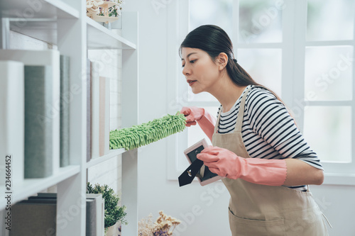 Canvas Print asian housekeeper in apron dusting the bookshelf