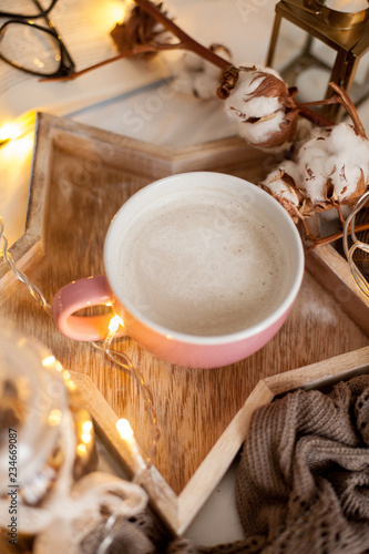 Mug of hot cappuccino on a wooden tray is on the bed. Cozy decor. Breakfast. Mug, plaid, cotton, candle. Book. Christmas lights. Holidays. Christmas. Autumn. Winter.
