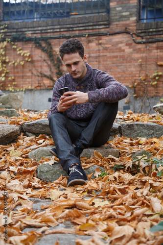 Young handsome man sitting on a rock in meadow with foliage and speaking with smartphone