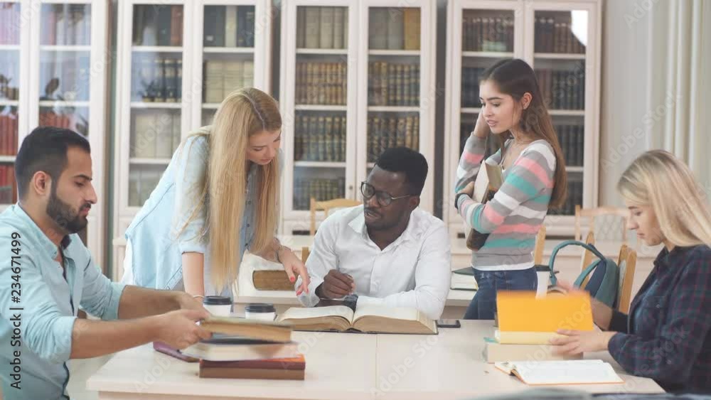 Multiethnic young people sitting at table reading big old reference book for preparing scientific report. Group of young students doing together homework at library. Multiethnic young people sitting at table reading big old reference book for preparing scientific report. Group of young students doing together homework at library.