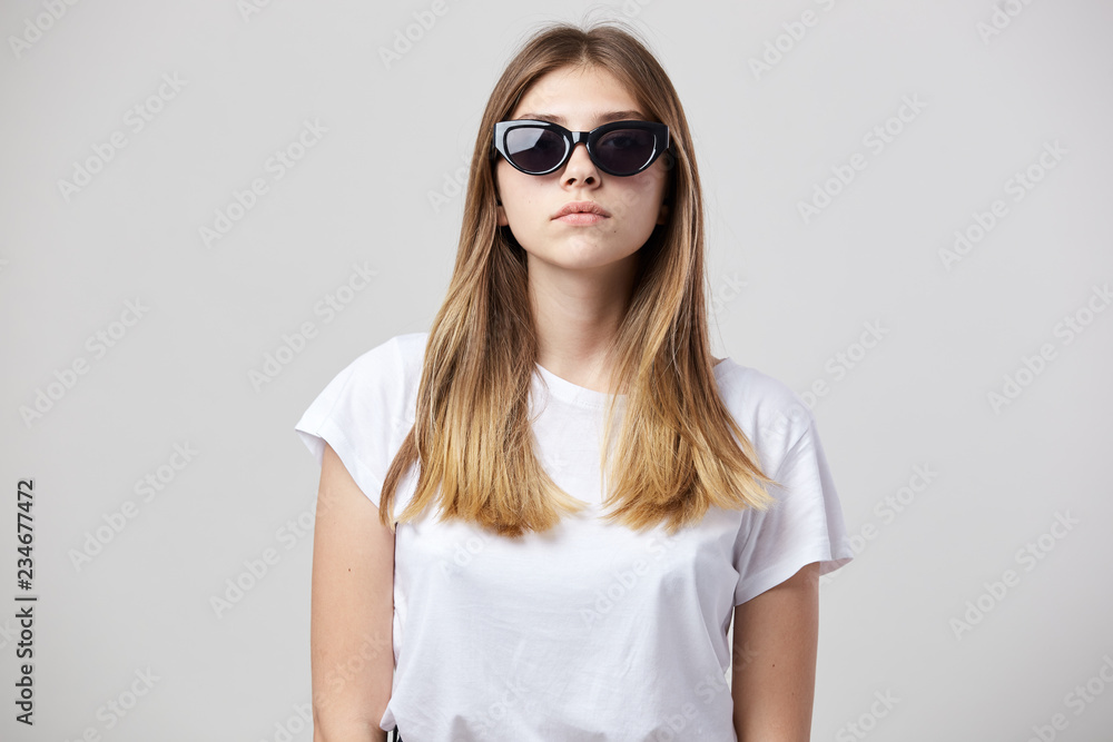 Serious girl dressed in a white t-shirt and sun glasses is on a white background in the studio Serious girl dressed in a white t-shirt and sun glasses is on a white background in the studio