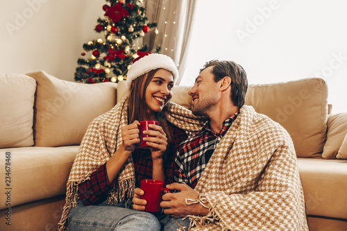 Christmas. Love. Home. Young couple in Santa hats, covered in plaid is holding cups, talking and smiling while sitting at home near the Christmas tree photo