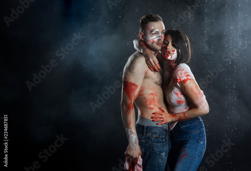 Couple of artists wearing jeans and t-shirts soiled with paint hug each other under rain in smoke on black background. Palm prints and paints on their faces. Man with naked torso