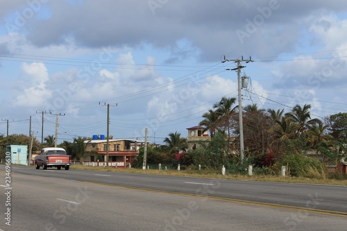 Vintage American car driving down the highway between Varadero and Havana, Cuba. There are houses and other buildings alongside the road, as well as palm trees and other tropical trees and plants. The